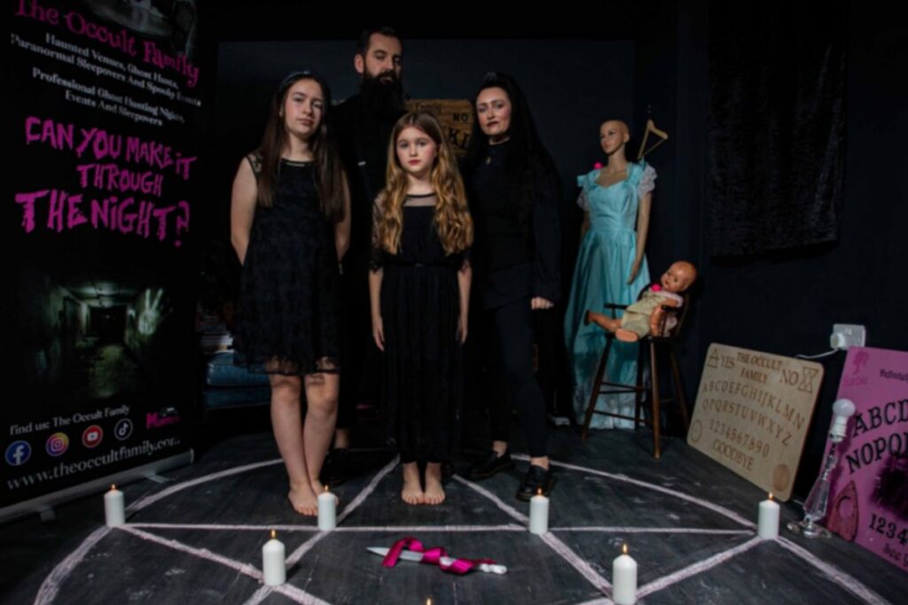 Perhe seisoo ouijalautojen keskellä pentagrammin päällä mustassa huoneessa. Perhe on pukeutunut Addams Family henkisiin vaatteisiin. 