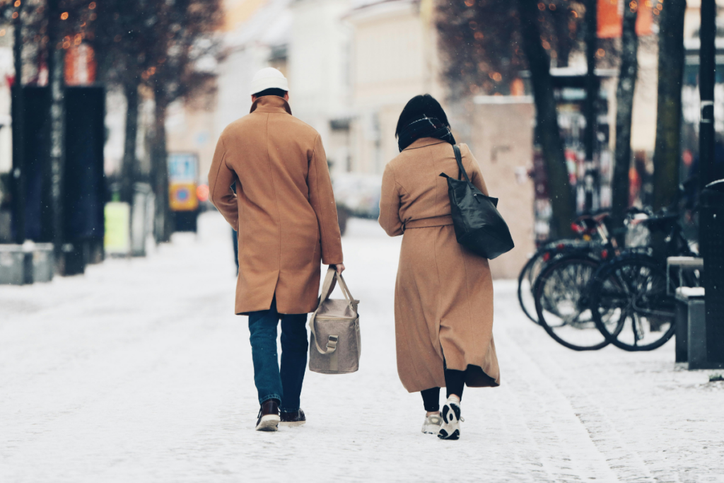 Kaksi aikuista kävelee poispäin kamerasta lumisella kaupunkikadulla. Molemmilla on pitkät ruskeat takit; toisella kädessä kangaskassi ja toisella olalla laukku. Taustalla näkyy polkupyöriä ja talvisia kaupunkirakennuksia.