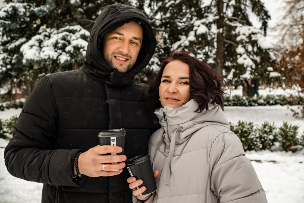 Mies ja nainen seisovat ulkona lumisessa puistossa talvivaatteissa ja pitävät käsissään kahvikuppeja, taustalla lumisia puita ja pensaita.