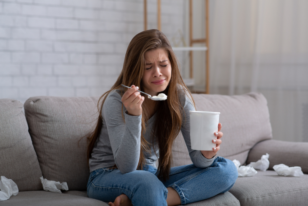 Nainen istuu sohvalla harmaassa paidassa ja sinisissä farkuissa, itkuisena ja surullisena. Hän pitelee isoa jäätelörasiaa ja lusikkaa, ympärillä on rutistettuja nenäliinoja. Kuvassa näkyy eron jälkeinen suru ja lohdun etsiminen arjen pienistä asioista.