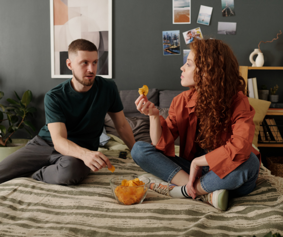 Kuvassa mies ja nainen syövät sängyllä sipsejä ja juttelevat.