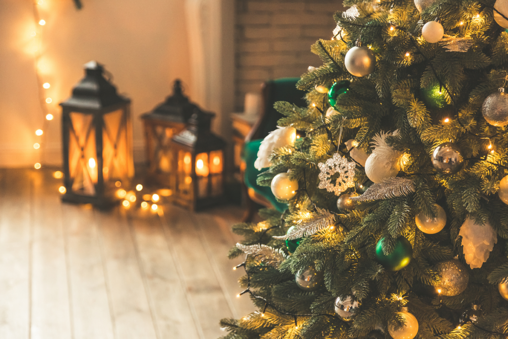 Sisätiloissa koristeltu joulukuusi, jossa lämpimän sävyiset valot ja hopean- ja vihreänsävyisiä joulupalloja, taustalla lattialle asetettuja lyhtyjä ja kynttilänvaloa.