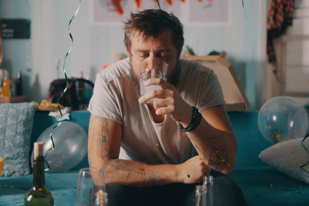 Parrakas mies istuu sohvalla juhlan jälkeisessä ympäristössä ja juo vettä lasista silmät suljettuina. Pöydällä ja lattialla näkyy tyhjiä pulloja, kertakäyttöastioita ja koristeita, jotka viittaavat krapulaan juhlien jälkeen.