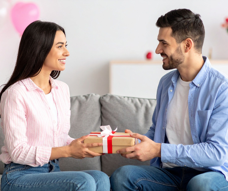 Kuvassa mies antaa naiselle lahjan sohvalla hymyillen. Taustalla näkyy vaaleanpunaisia ilmapalloja