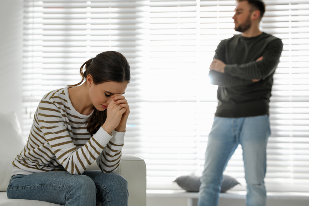Nainen istuu nojatuolilla kädet yhteen puristettuina ja pää painettuna alas, selvästi ahdistuneena. Taustalla mies seisoo kädet ristissä ja katsoo sivulle, etäisenä ja sulkeutuneena. Kuva symboloi parisuhteen kriisiä, pettymyksen tai erimielisyyden hetkeä.