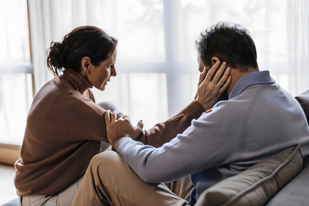 Nainen istuu sohvalla miehen edessä ja koskettaa häntä olkapäältä ja kasvoilta, mies peittää osittain kasvonsa kädellään ja kuuntelee, ympärillä pehmeä luonnonvalo.