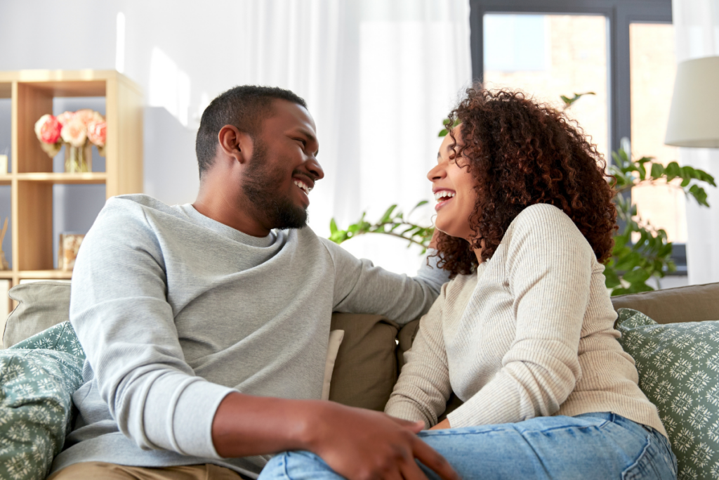 Pariskunta istuu sohvalla valoisassa olohuoneessa, nauraa ja katsoo toisiaan iloisesti. Takana näkyy viherkasveja ja kirjahylly, ja huoneen sisustus luo kodikkaan ja läheisen tunnelman.