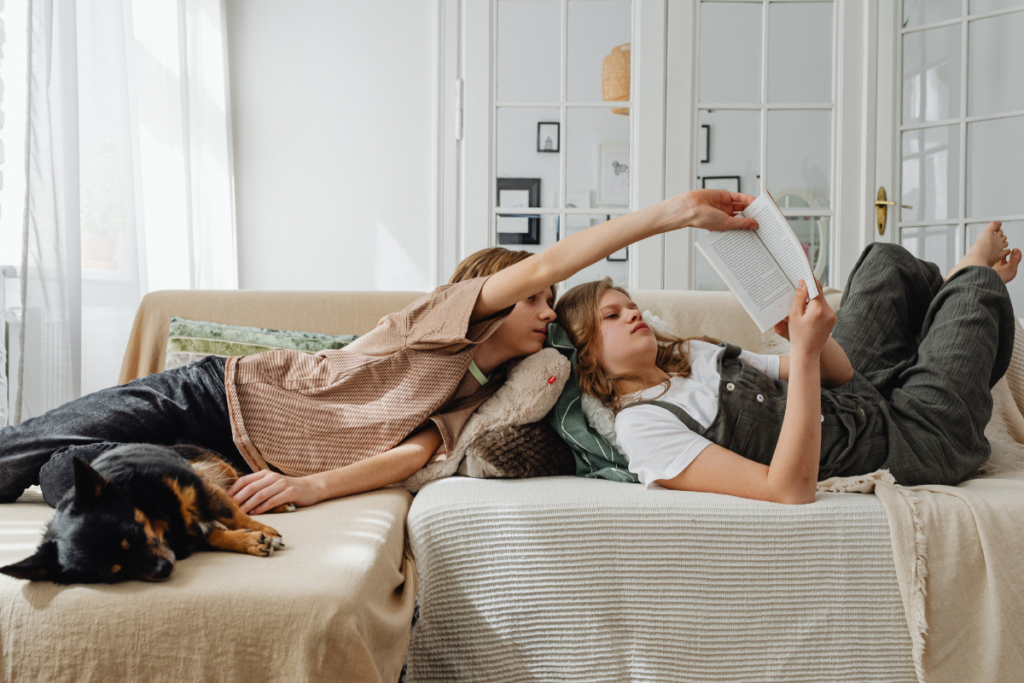 Kaksi lasta makaa rennosti vaalealla sohvalla olohuoneessa ja lukee kirjaa yhdessä. Toinen lapsista pitelee kirjaa ylhäällä, kun toinen nojaa vieressä ja katsoo sivuja. Sohvan vieressä nukkuu mustaruskea koira, ja huoneessa on rauhallinen, kodikas tunnelma.