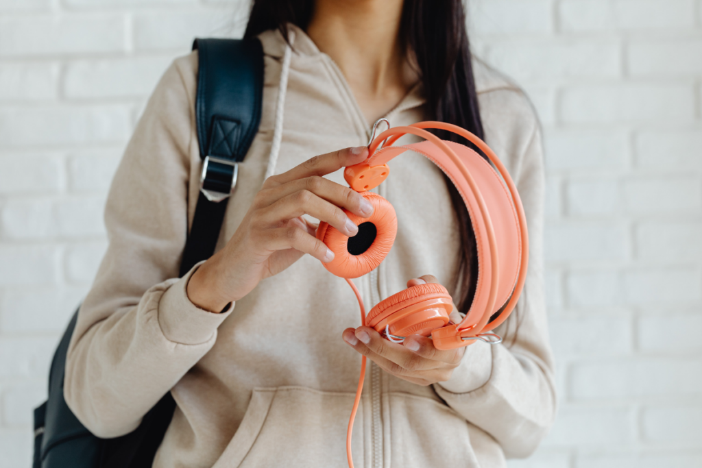 Henkilö pitää käsissään oransseja langallisia kuulokkeita, taustalla vaalea tiiliseinä ja olalla reppu.