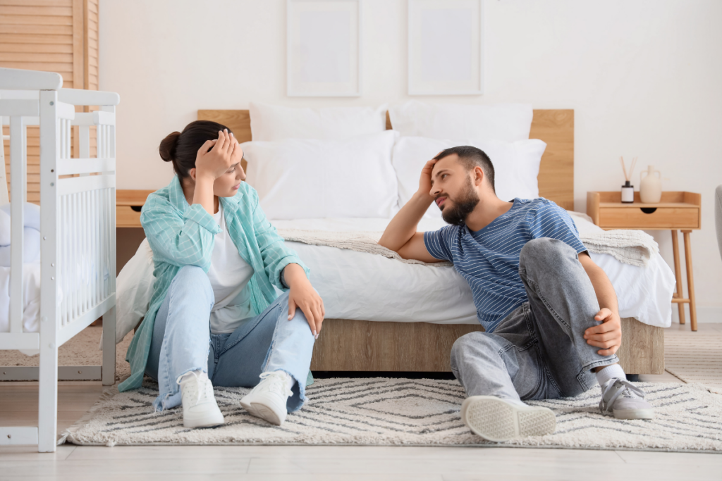 Kuvassa vanhemmat istuvat lastenhuoneen lattialla väsyneinä ja uupuneina ja keskustevat keskenään.