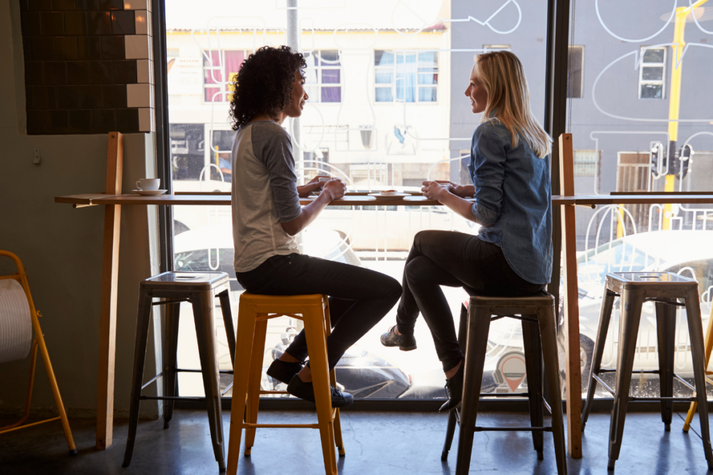 Kaksi naista istuu vastakkain korkeilla jakkaroilla ikkunan ääressä kahvilassa ja keskustelee, pöydällä on kupit ja ikkunasta näkyy katumaisema.