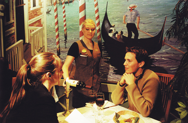 Ravintola Omenapuun tarjoilija kaataa viiniä pöydässä istuvalle pariskunnalle ravintolassa. Asiakkaat keskustelevat pöydän ääressä, jolla on viinilasit ja leipäkori. Taustalla on seinämaalaus Venetsian kanavasta gondoleineen, mikä luo klassisen ja romanttisen ruokailutunnelman.