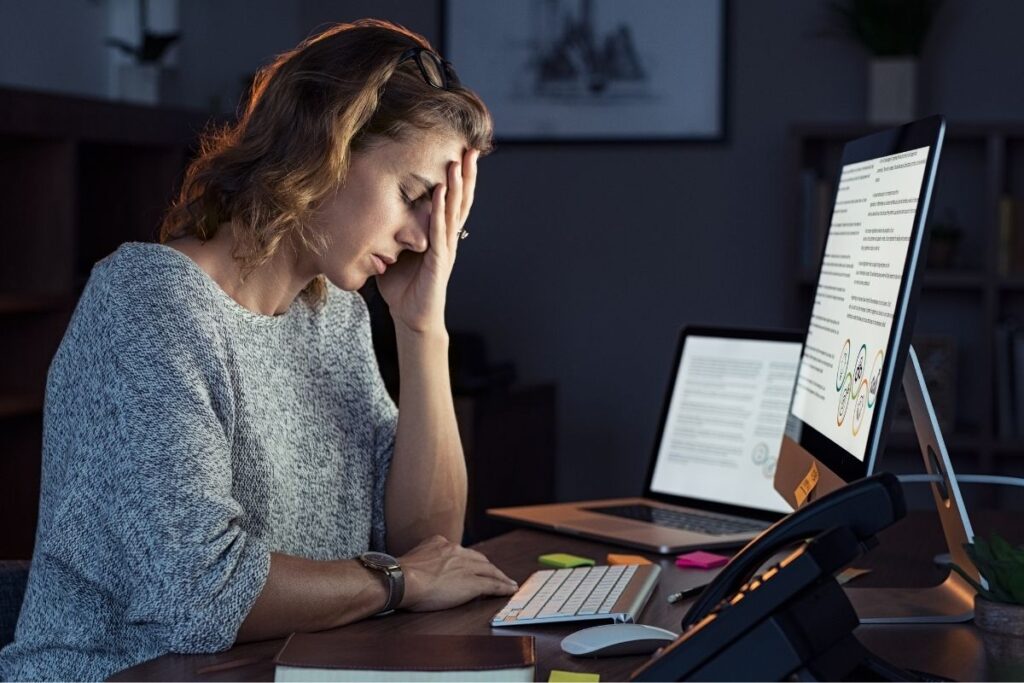 Nainen istuu työpisteellään ja vaikuttaa uupuneelta. Hän nojaa käteensä silmät kiinni. 
