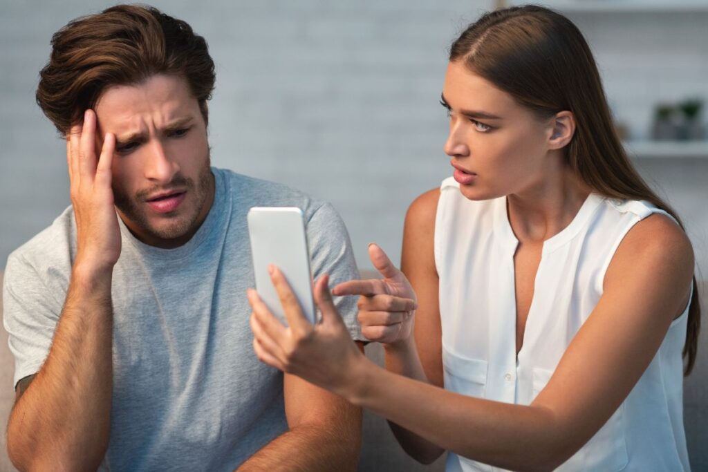 Mies ja nainen katsovat puhelimen näyttöä. Nainen on selkeästi löytänyt jotain miehen puhelimesta ja mies näyttää tuskastuneelta ja kiinni jääneeltä. He istuvat kodinomaisessa ympäristössä.