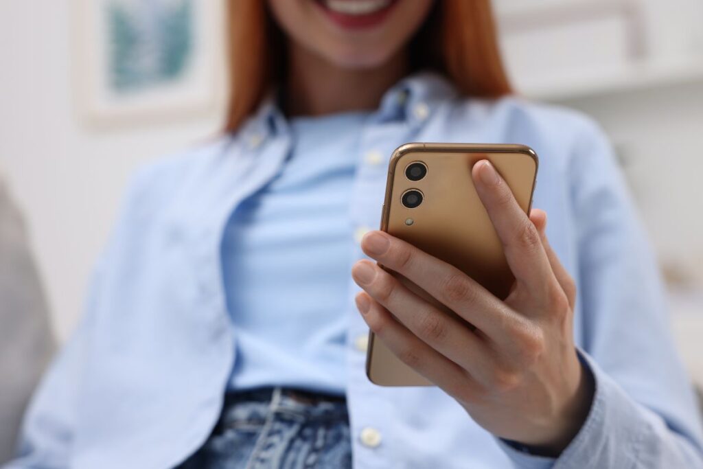 Sinipaitainen nainen hymyilee sille, mitä näkee puhelimensa ruudulta. Hänestä ei näy kuin leuka ja hampaat. 