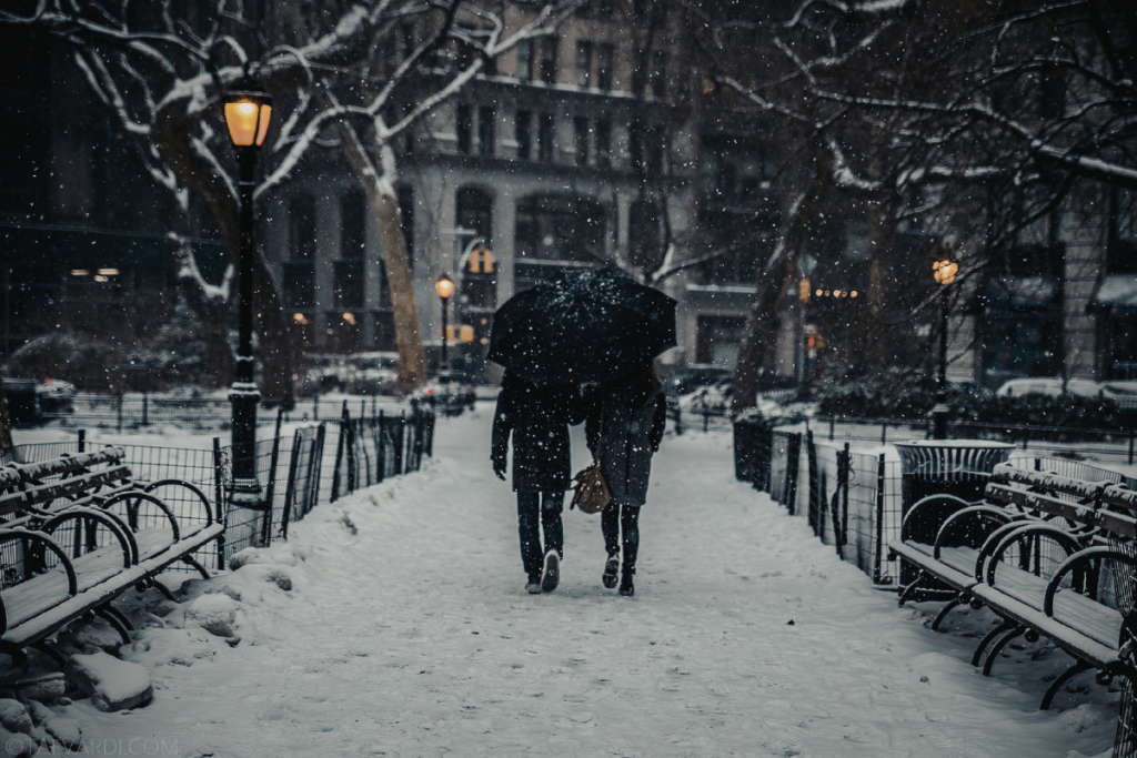 Pariskunta kävelee käsi kädessä lumisessa kaupunkipuistossa iltahämärässä, jakaa sateenvarjon ja etenee penkkien reunustamaa polkua pitkin.
