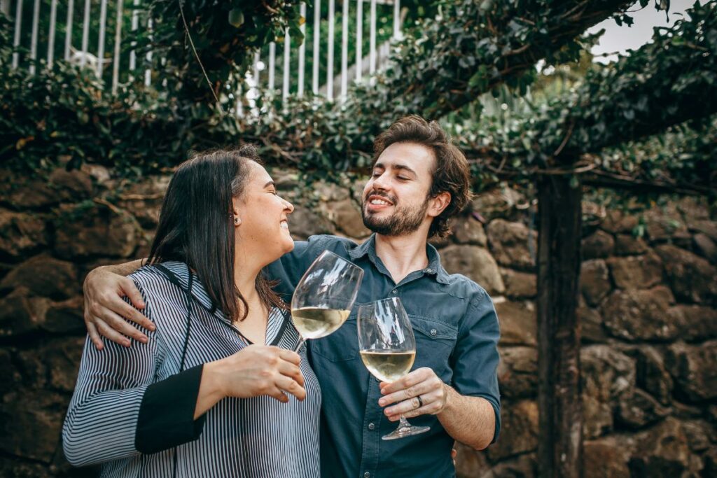 Pariskunta seisoo lähekkäin valkoviinilasit käsissään ja he hymyilevät toisilleen. He seisovat kasvien peitossa olevan katoksen alla, ja kippistelevät.