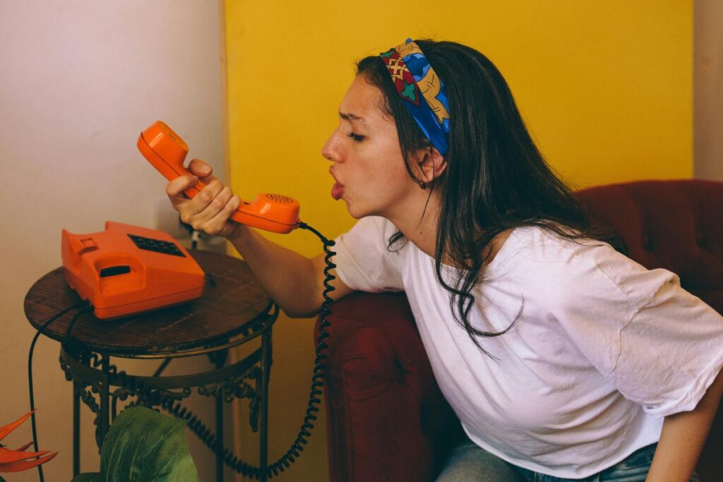 Nuori nainen istuu sisätiloissa ja puhuu oranssiin lankapuhelimeen, taustalla värikäs seinä ja huonekalu.