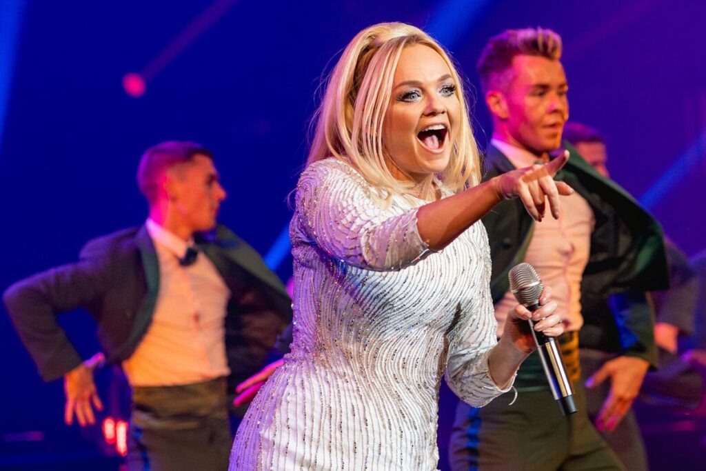 Vaaleahiuksinen naisartisti esiintyy lavalla mikrofonin kanssa ja osoittaa yleisöä, taustalla tanssijoita värikkäissä lavavaloissa.