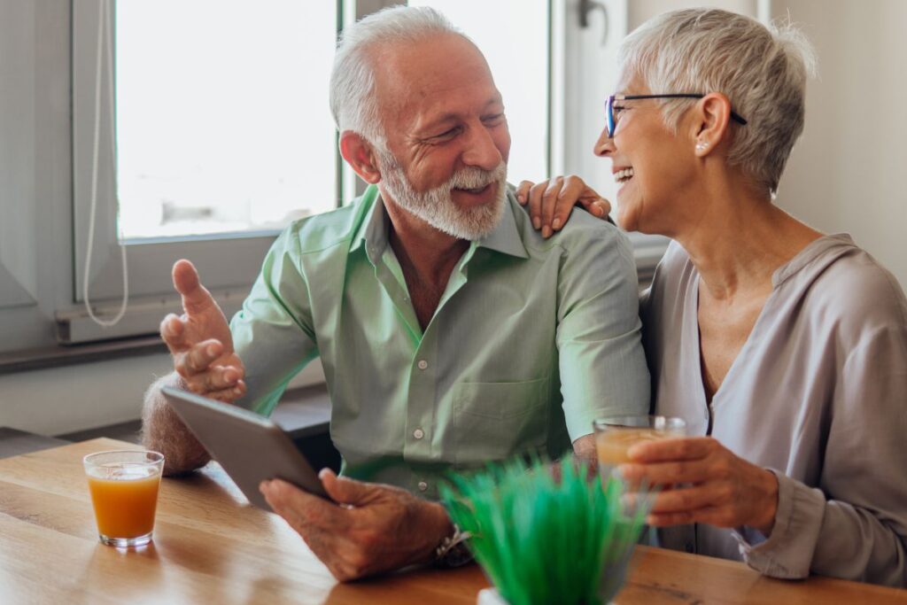 Iäkäs mies ja nainen istuvat pöydän ääressä, keskustelevat ja hymyilevät toisiaan katsoen, miehen kädessä on tablettilaite ja molemmilla juomalasit edessään.