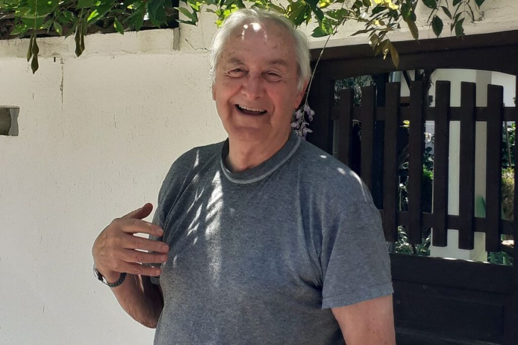 Iäkäs mies hymyilee kameralle kiviaidan vieressä harmaassa t-paidassa. 