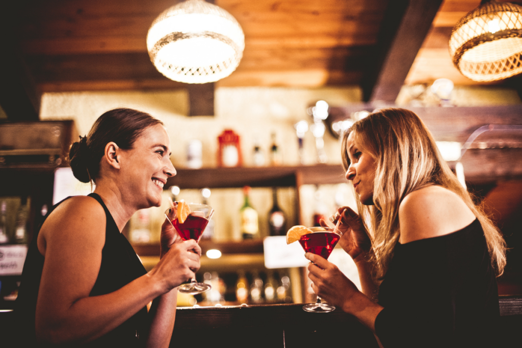 Kaksi ihmistä istuu vastakkain baarissa ja hymyilee toisilleen pitäen käsissään punaisia cocktaileja, joissa on appelsiiniviipale koristeena.