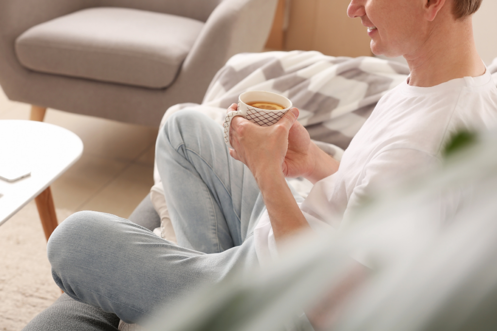 Henkilö istuu sohvalla vaaleassa olohuoneessa ja pitää käsissään kahvikuppia, rennosti jalat ristissä.