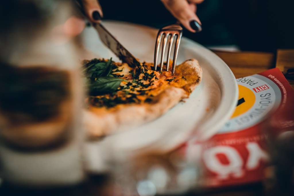 Lähikuva lautasella olevasta pizzasta, jota leikataan veitsellä ja haarukalla; pöydällä näkyy juomalasi ja osittain Syö!-viikot-kampanjan materiaalia.