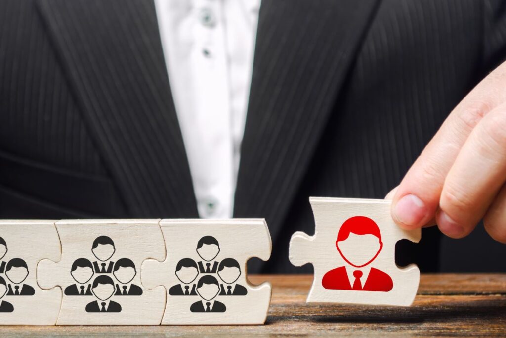 Käsi nostaa palapelin palaa, jossa on yhden henkilön kuvake, muiden palojen esittäessä ryhmiä ihmishahmoja pöydällä.