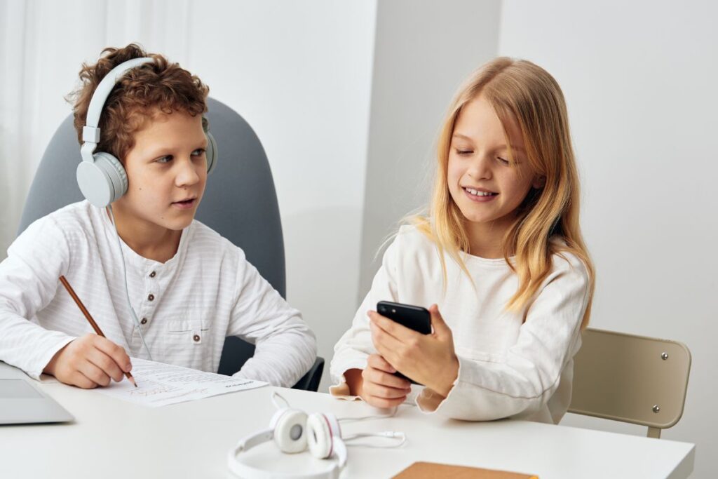 Kaksi lasta istuu pöydän ääressä, toinen katsoo älypuhelinta ja toinen tekee tehtäviä kuulokkeet päässä, pöydällä on kuulokkeet.