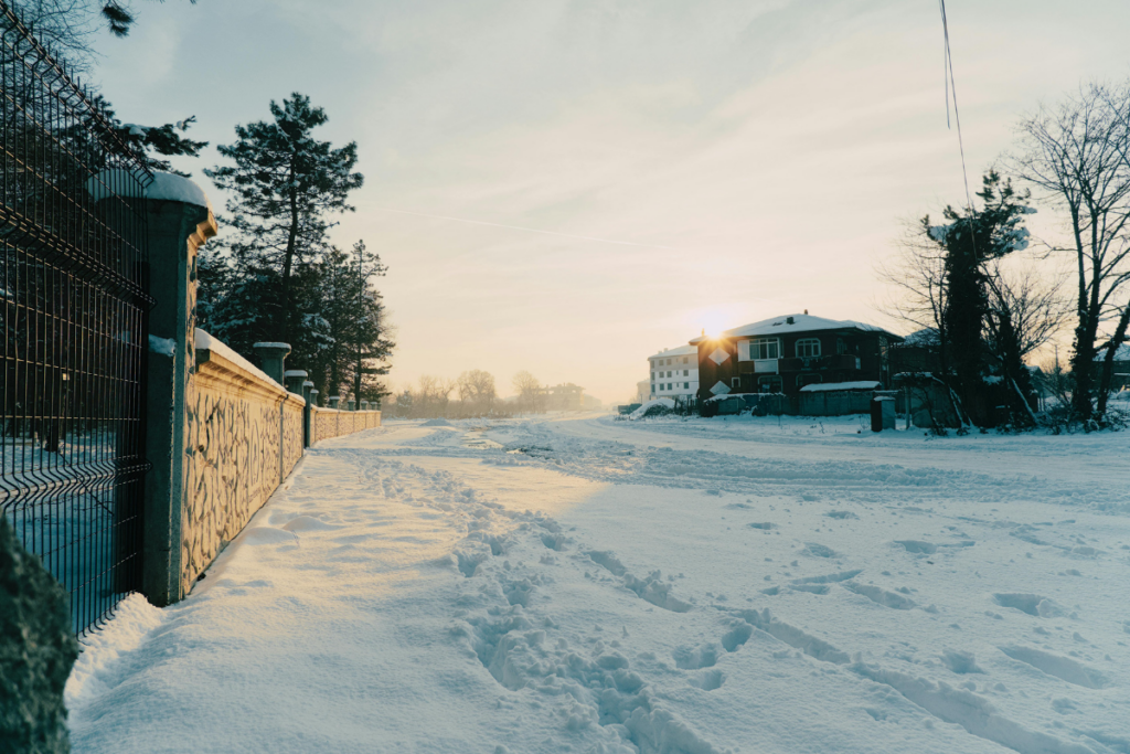 Talvinen asuinkatu varhain aamulla; lumi peittää tien ja rakennukset, ja matala aurinko nousee horisontissa valaisten maisemaa.