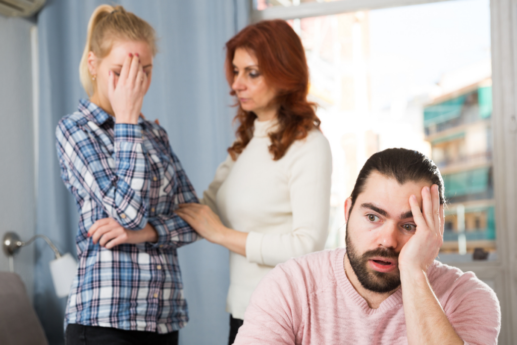 Kolme henkilöä huoneessa. Etualalla mies istuu käsi otsalla, taustalla nainen koskettaa toisen naisen olkapäätä, joka seisoo silmät peittäen.