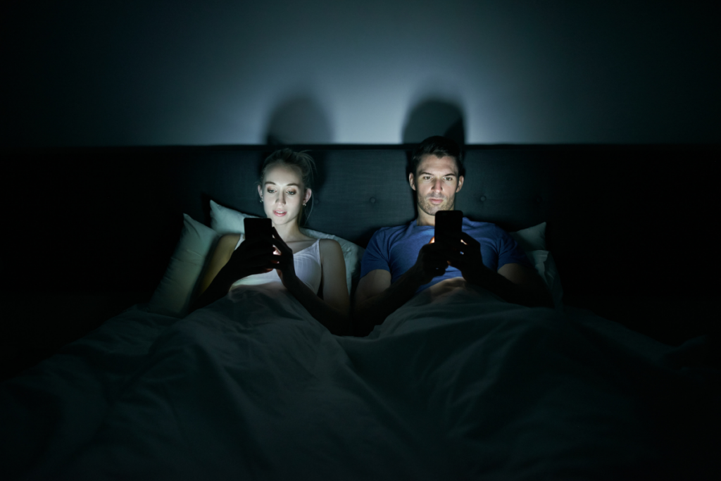Pariskunta makaa sängyssä pimeässä huoneessa ja katsoo puhelimiaan, ruutujen valo valaisee heidän kasvonsa.