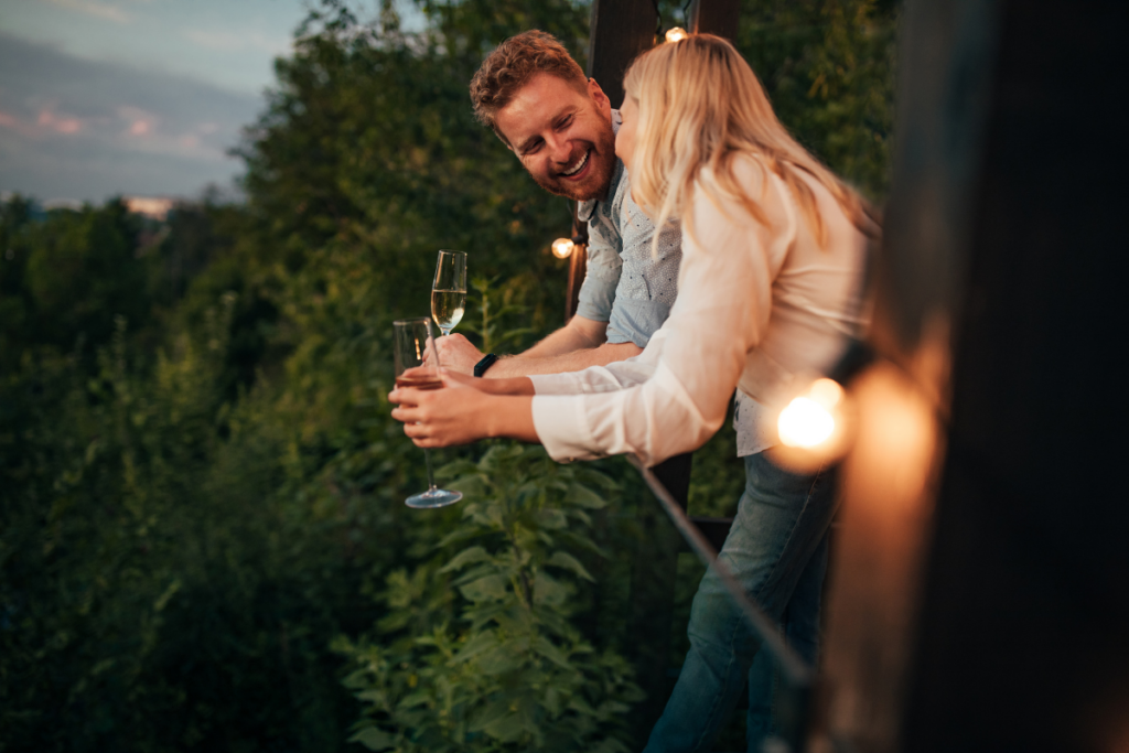 Hymyilevä pariskunta seisoo ulkona parvekkeella tai terassilla iltavalaistuksessa, pitelee kuohuviinilaseja ja nojaa kaiteeseen vehreän maiseman yläpuolella.