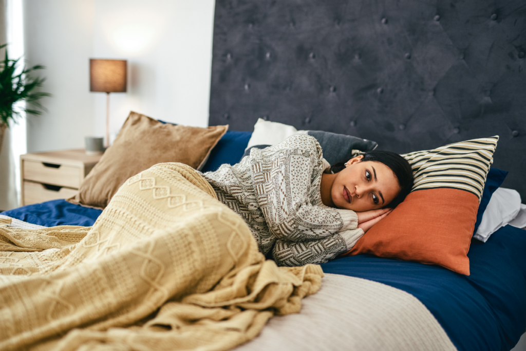 Nainen makaa yksin sängyssä peiton alla kyljellään ja katsoo ajatuksiinsa vaipuneena eteenpäin makuuhuoneessa.