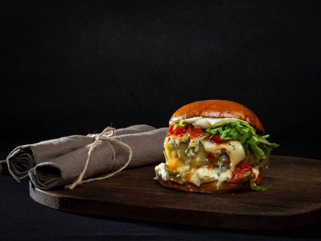 Hampurilainen briossisämpylässä, sulanutta juustoa, salaattia ja kastiketta puualustalla.