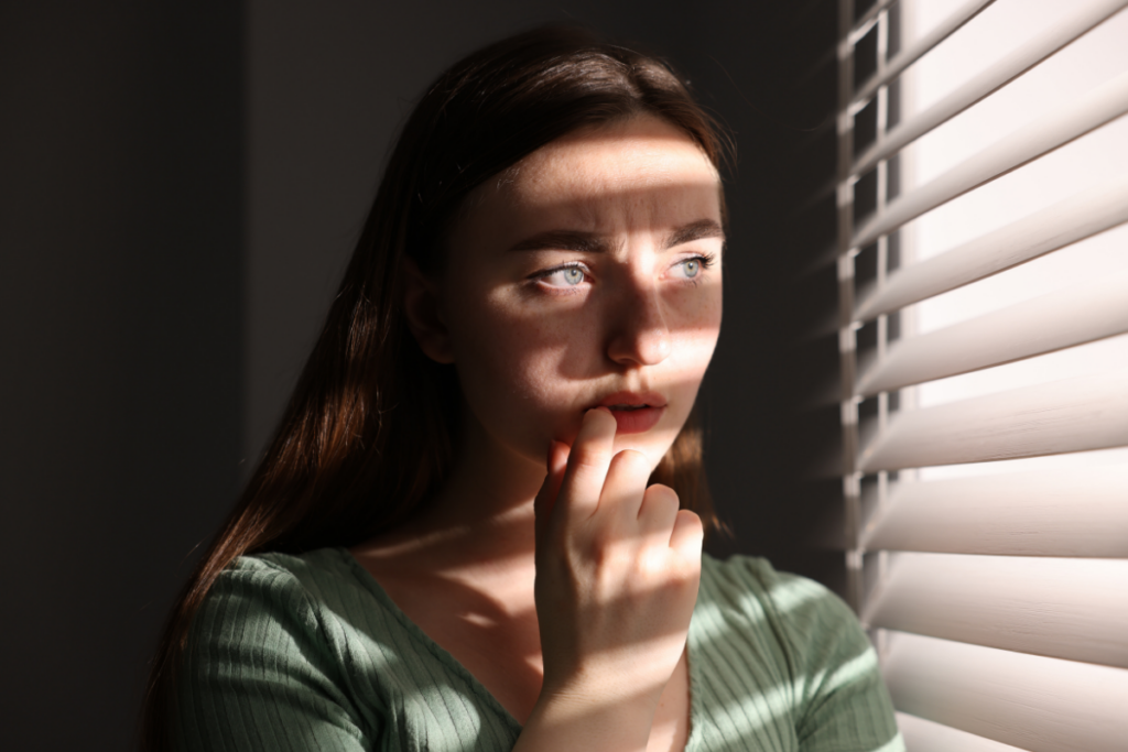 Nuori nainen seisoo ikkunan vieressä ja katsoo ulos sälekaihtimien läpi. Valon ja varjojen raidat osuvat hänen kasvoilleen, ja hän koskettaa huultaan mietteliäänä, ilme huolestunut ja pohdiskeleva.