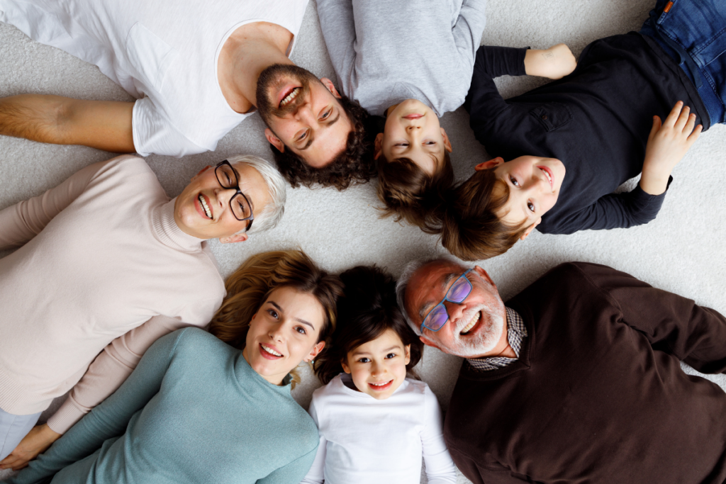 Usean sukupolven perhe makaa lattialla päät vastakkain ympyrän muodossa. Isovanhemmat, aikuiset ja lapset katsovat ylöspäin kameraan ja hymyilevät, muodostaen läheisen ja iloisen perhepotretin.
