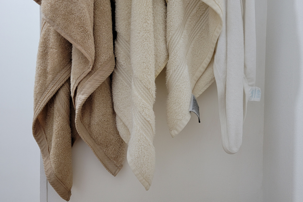 Useita erivärisiä kylpypyyhkeitä roikkumassa vierekkäin kylpyhuoneen seinäkoukuissa, vaalea seinä ja rauhallinen valaistus taustalla.