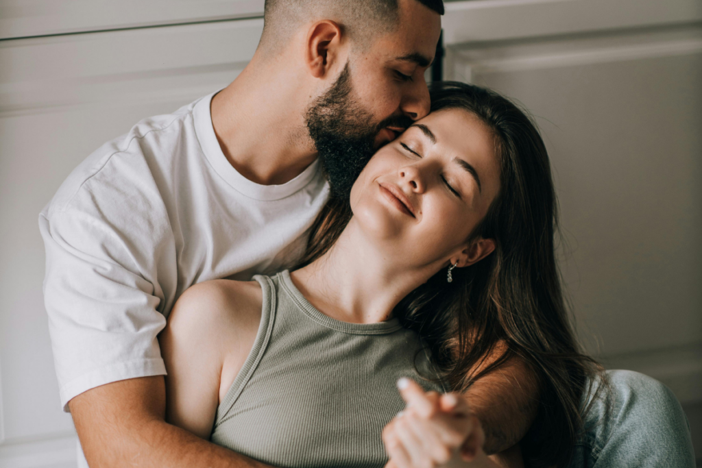 Mies pitää naista sylissään ja suutelee tätä hellästi ohimolle, nainen nojaa taaksepäin silmät kiinni ja hymyilee rauhallisesti, hetki kuvastaa turvallisuutta, kiintymystä ja emotionaalista läheisyyttä.