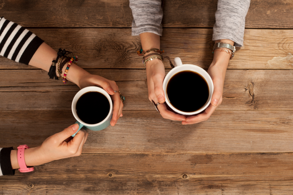 Ylhäältä päin kuvattu näkymä kahdesta ihmisestä, jotka pitelevät kahvikuppeja puupöydän ääressä. Molemmissa kupeissa on mustaa kahvia, ja käsissä näkyy rannekoruja ja kelloja.