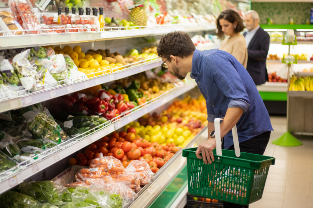 Mies valitsee vihanneksia supermarketin hedelmä- ja vihannesosastolla ja pitää kädessään vihreää ostoskoria; taustalla kaksi muuta asiakasta selaa tuotteita värikkäiden hyllyjen äärellä.