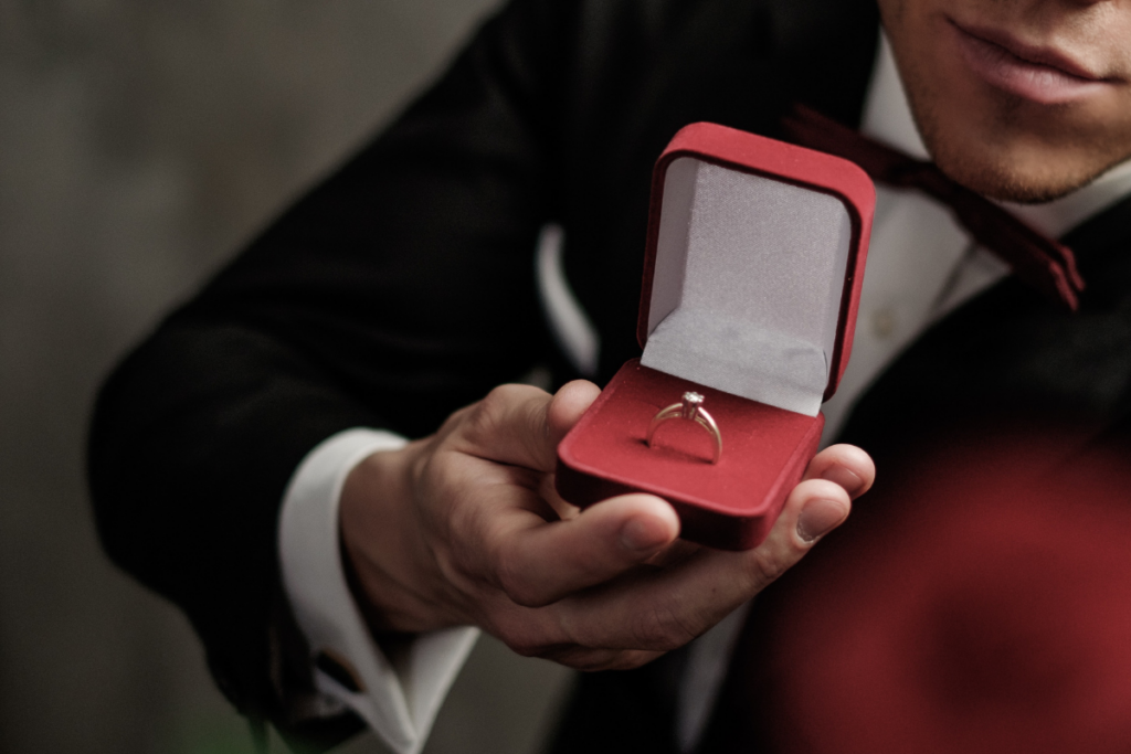 Mies pujottaa sormusta toisen henkilön nimettömään pöydän ääressä, ja molempien kädet ovat kuvan keskiössä. Tilanne kuvaa intiimiä kosinta- tai kihlautumishetkeä, jossa ele on rauhallinen ja tunteikas.