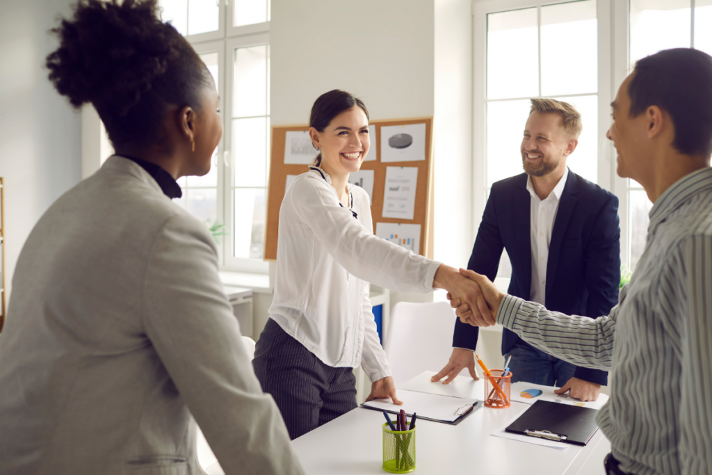Neljän hengen työryhmä seisoo valoisassa toimistossa pöydän äärellä; keskellä oleva nainen kättelee miestä, ja muut tiimin jäsenet seuraavat tilannetta hymyillen, pöydällä näkyy asiakirjoja ja toimistotarvikkeita.