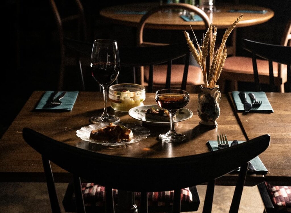 Kattaus hämärässä ravintolassa: puupöytä, viinilasit, pienet annokset ja kynttilänvalo.