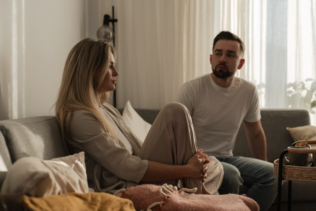 Nainen istuu sohvalla polvet vedettynä rintaansa vasten ja katsoo poispäin miehestä, joka istuu vieressä ja katsoo häntä huolestuneena; tunnelma on vakava ja etäinen, viitaten jännitteeseen tai ristiriitaan parisuhteessa.