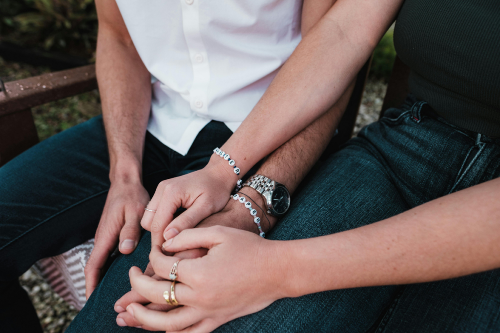 Lähikuva kahden ihmisen käsistä heidän istuessaan vierekkäin penkillä ulkona. He pitävät toisiaan kädestä; ranteissa näkyy yhteensopivia rannekoruja ja sormissa sormuksia. Kuvassa korostuu läheisyys ja yhteenkuuluvuus.