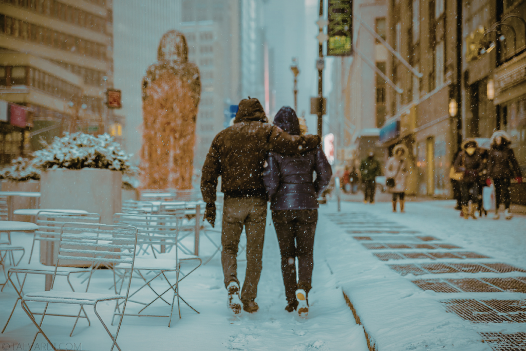 Pariskunta kävelee käsi kädessä lumisella kaupunkikadulla, toinen pitää kättään toisen hartioilla, ympärillä sataa lunta ja taustalla näkyy ihmisiä ja rakennuksia.