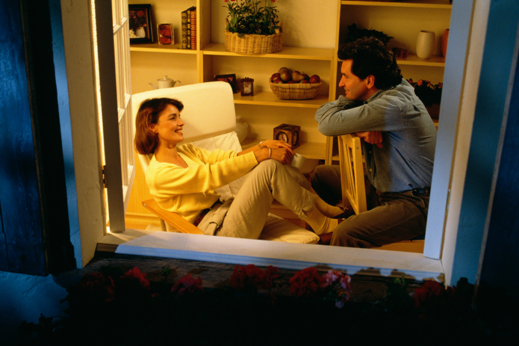 Onnellinen pari istuu vastakkain kodikkaassa olohuoneessa iltahämärässä. He juttelevat ja hymyilevät toisilleen lämpimässä valossa, ympärillä hyllyjä ja kodin yksityiskohtia, jotka korostavat rauhallista ja intiimiä tunnelmaa.