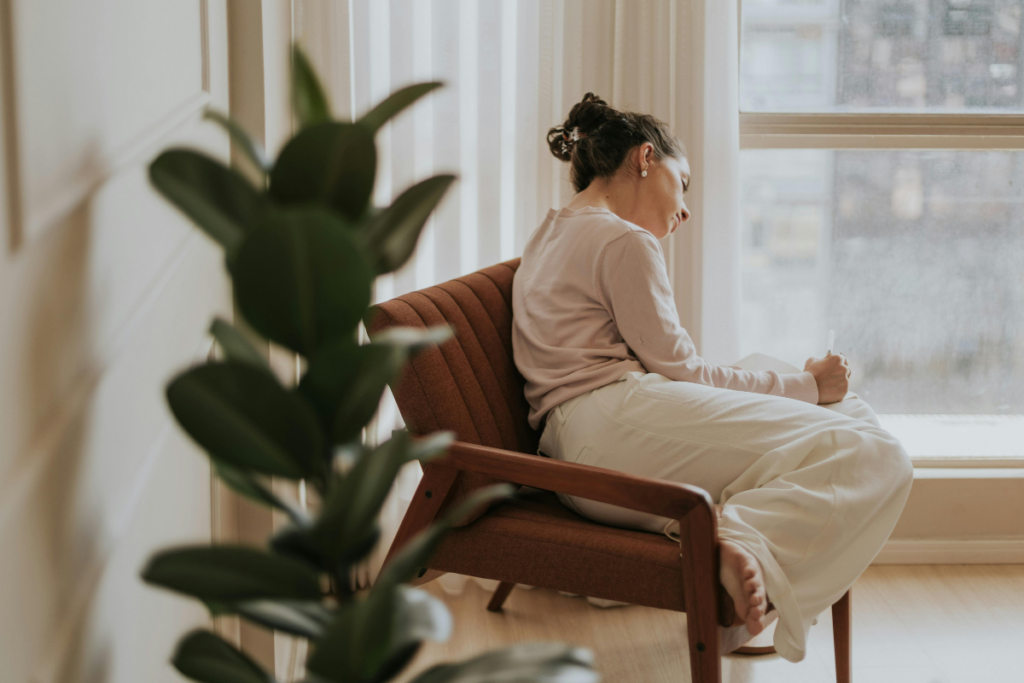 Nainen istuu nojatuolissa ikkunan vieressä jalat tuolilla käpertyneinä ja katsoo alaspäin kädessään olevaa mukia, kun pehmeä päivänvalo ja huonekasvi etualalla luovat hiljaisen ja introverttia rauhaa huokuvan tunnelman.